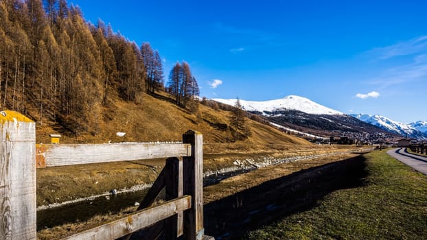 Beautiful mountain valley with stream, trees, wooden fence and footpath, Livingo village in background, Italy, Alps