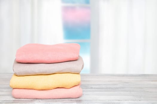 Stack of winter clothes. Pile of colorful knitted cozy warm cashmere sweaters or pullover on wooden table against abstract bright winter background. Space for display product montage. Beautiful backdrop.