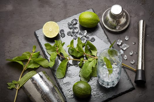 Set of bar tools: cocktail shaker, muddler and glass with ice, lime and mint on dark stone table. Top view. Bartender's tool. Mojito cocktail making.