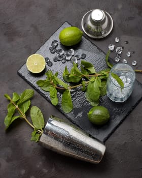 bar tool: cocktail shaker and glass with ice, lime and mint on dark stone table. Top view. Bartender's tool. Mojito cocktail making.