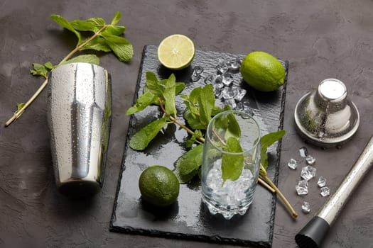 Set of bar tools: cocktail shaker, muddler and glass with ice, lime and mint on dark stone table. Top view. Bartender's tool. Mojito cocktail making.