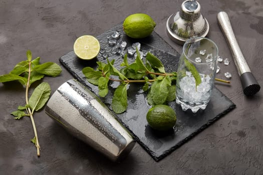 Set of bar tools: cocktail shaker, muddler and glass with ice, lime and mint on dark stone table. Top view. Bartender's tool. Mojito cocktail making.
