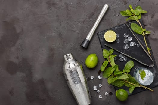 Set of bar tools: cocktail shaker, muddler, ice tongs and glass with ice, lime and mint on dark stone table. Top view with copy space. Bartender's tool. Mojito cocktail making.