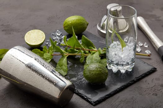 Set of bar tools: cocktail shaker, muddler and glass with ice, lime and mint on dark stone table. Top view. Bartender's tool. Mojito cocktail making.