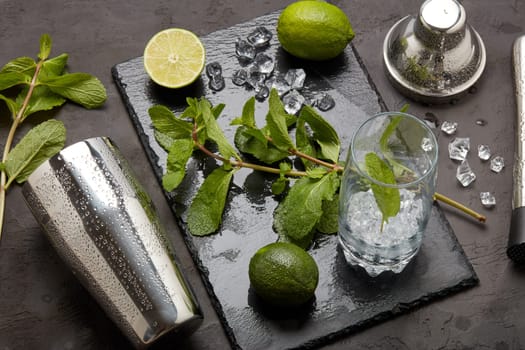 Set of bar tools: cocktail shaker, muddler and glass with ice, lime and mint on dark stone table. Top view. Bartender's tool. Mojito cocktail making.