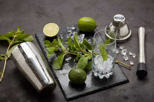 Set of bar tools: cocktail shaker, muddler and glass with ice, lime and mint on dark stone table. Top view. Bartender's tool. Mojito cocktail making.