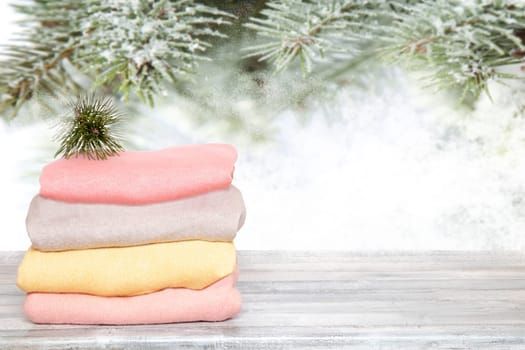 Pile of colorful knitted cozy warm cashmere sweaters on wooden table against blurred abstract snowy fir branches xmas background. Template for christmas sale.