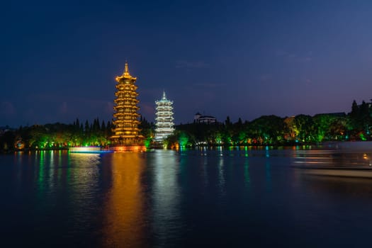 Tourist boats sailing in front of illuminated at night Sun and Moon Pagodas Towers at Shanhu or Shan Lake in Guilin town, Guangxi Province, China
