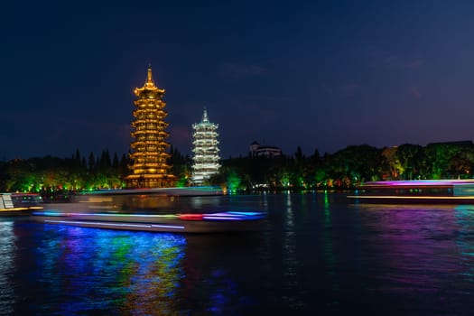 Tourist boats sailing in front of illuminated at night Sun and Moon Pagodas Towers at Shanhu or Shan Lake in Guilin town, Guangxi Province, China
