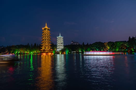 Tourist boats sailing in front of illuminated at night Sun and Moon Pagodas Towers at Shanhu or Shan Lake in Guilin town, Guangxi Province, China