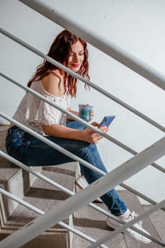 Young woman with red hair sitting on stairs and speaking on her smartphone and drinking coffee.