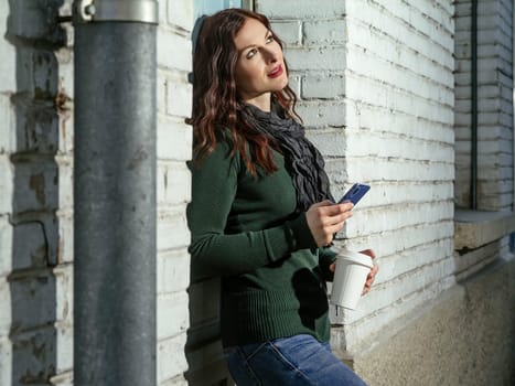 Young woman with red hair using her cellphone and drinking coffee from a takeout cup.