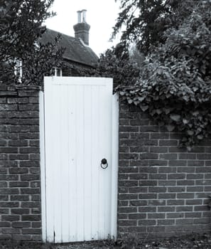 White Garden Gate in a brick wall leading to an English cottage in monochrome. With Chimney and trees showing in background