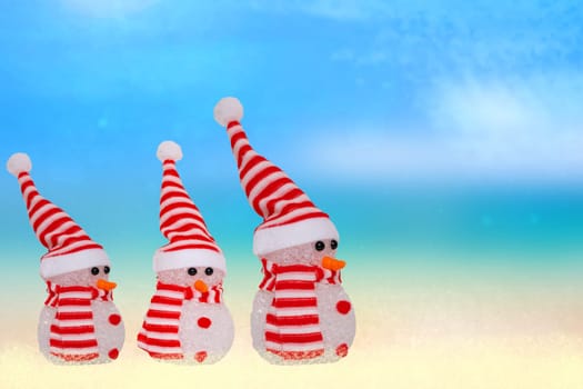 Tropical christmas background. A snowmen family with red white striped hat and scarf at astract tropical sandy beach with blue sky, clouds and ocean. Concept xmas holiday in hot countries.