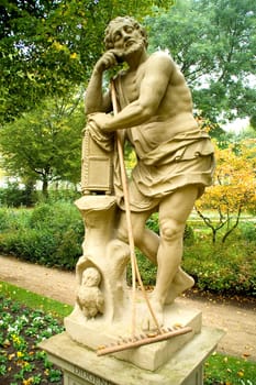 Diogenes sculpture in a relaxed position in a parc with a rake leaning unused in his arms.