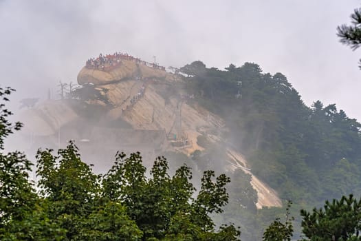 Huashan, China -  August 2019 : Crowds of tourists standing on a scenic viewing lookout platform on top of a narrow ridge on a mountain trail to the West Peak on Huashan mountain, Shaanxi Province