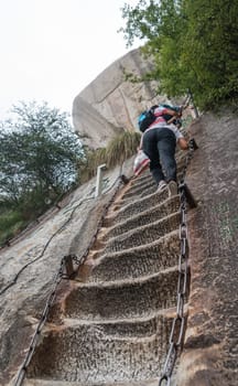 Huashan, China -  August 2019 : Tourist with backpack climbing up the steep, cut into rocky edge stairs on a mountain trail to the North and West Peak on Huashan mountain, Xian, Shaanxi Province