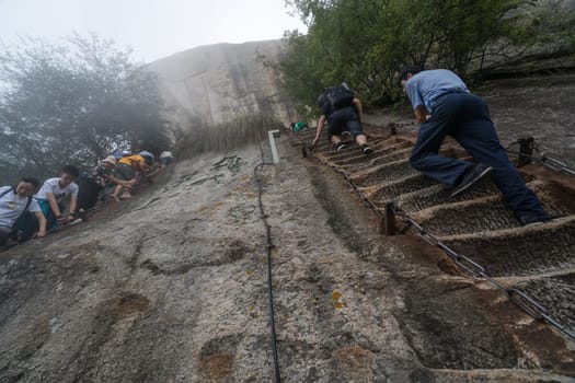 Huashan, China -  August 2019 : Tourists climbing up the vertical, steep, cut into rocky edge stairs on a mountain trail to the North and West Peak on Huashan mountain, Xian, Shaanxi Province