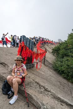 Huashan, China -  August 2019 : Tired chinese woman with a walking pole sitting on a rocky ground and resting on a mountain trail to the West Peak on Huashan mountain, Xian, Shaanxi Province