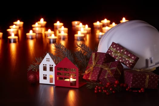 White construction hard hat, candles, gift boxes, fir branch and Christmas ornament on black background. Construction New Year, Christmas background. For greeting card or advertising