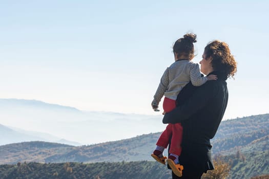 Young mother with little daughter looking at mountains on vacation, family travel together on top of high mountain outdoors, looking at beautiful landscape. Rear viewpoint