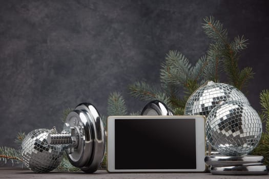 Sports equipment (dumbbell with weights), fir branch, Christmas ornament, blank computer tablet on dark grey background with copy space. Fitness, sport, healthy lifestyle concept. New Year. Christmas