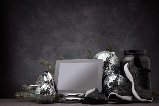 Sports nutrition, sports equipment, fir branch, Christmas ornament, blank computer tablet on dark grey background with copy space. Fitness, sport or healthy lifestyle concept. New Year and Christmas.