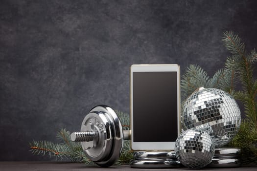 Sports equipment (dumbbell with weights), fir branch, Christmas ornament, blank computer tablet on dark grey background with copy space. Fitness, sport, healthy lifestyle concept. New Year. Christmas