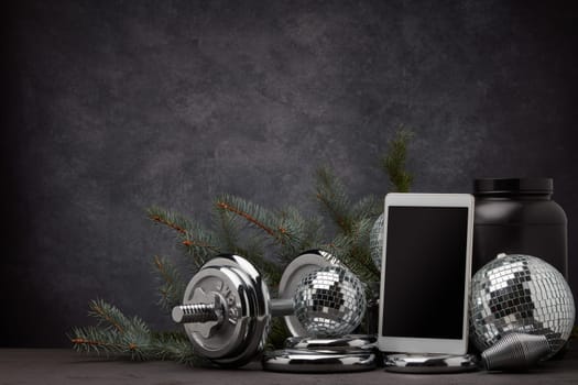 Sports nutrition, sports equipment, fir branch, Christmas ornament, blank computer tablet on dark grey background with copy space. Fitness, sport or healthy lifestyle concept. New Year and Christmas.