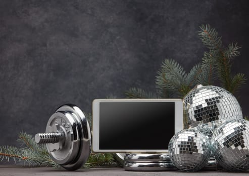 Sports equipment (dumbbell with weights), fir branch, Christmas ornament, blank computer tablet on dark grey background with copy space. Fitness, sport, healthy lifestyle concept. New Year. Christmas