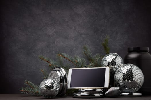 Sports nutrition, sports equipment, fir branch, Christmas ornament, blank computer tablet on dark grey background with copy space. Fitness, sport or healthy lifestyle concept. New Year and Christmas.