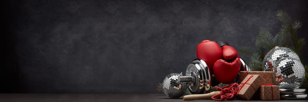 Sports equipment: dumbbell, boxing gloves, skipping rope, fir branch, Christmas ornament, gifts on dark grey background with copy space. Fitness, sport, healthy lifestyle concept. New Year. Christmas