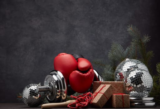 Sports equipment: dumbbell, boxing gloves, skipping rope, fir branch, Christmas ornament, gifts on dark grey background with copy space. Fitness, sport, healthy lifestyle concept. New Year. Christmas
