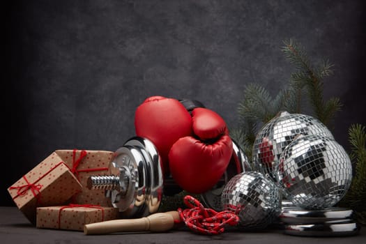 Sports equipment: dumbbell, boxing gloves, skipping rope, fir branch, Christmas ornament, gifts on dark grey background with copy space. Fitness, sport, healthy lifestyle concept. New Year. Christmas