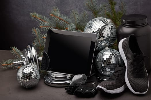 Sports nutrition, sports equipment, fir branch, Christmas ornament, blank computer tablet on dark grey background with copy space. Fitness, sport or healthy lifestyle concept. New Year and Christmas.