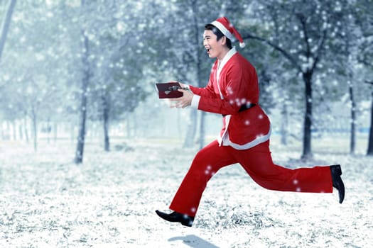 Asian man in Santa costume running while holding the gift box with snowy trees background