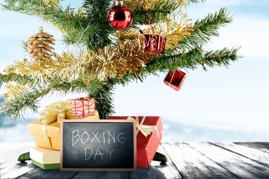 Christmas tree with gift box and Boxing Day text on the blackboard on wooden floor with a snowy background