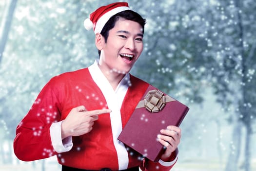 Asian man in Santa costume holding gift box with snowy trees background