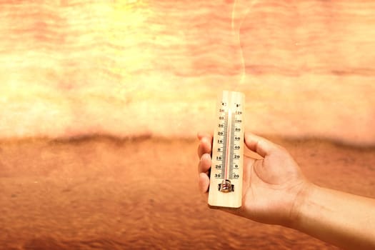 Hand holding thermometer with high temperature on the beach with glowing sun background. Heatwave concept
