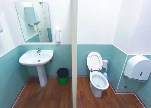 wide angle view of a toilet with the wooden floor and tiled wall with a mirror, a green basket, a sink and the ceramic bowl.