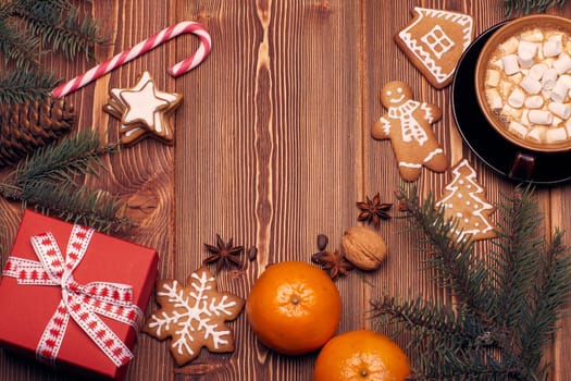 Christmas decor with fir tree branches, glass mug with hot cocoa and marshmallow, gingerbreads and gift box on a wooden background with copy space for text. Top view. For greeting card.