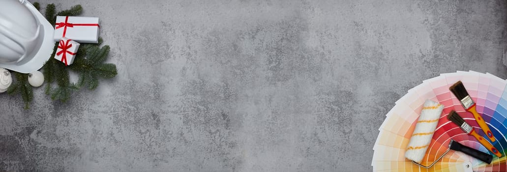 Construction hard hat, color palette guide, paint brushes and roller, fir tree branches and gift boxes on grey background. Top view with copy space, New Year and Christmas. Banner background