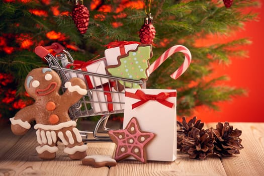 Shopping trolley with gift boxes and gingerbread cookies standing on a wooden table on a red background with Christmas fir tree. Christmas and New Year sale.