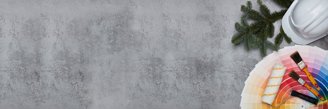 Construction hard hat, color palette guide, paint brushes and roller, fir tree branches on grey background. Top view with copy space, New Year and Christmas. Banner background