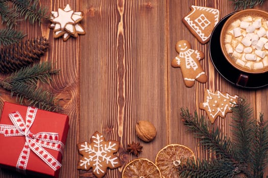 Christmas decor with fir tree branches, glass mug with hot cocoa and marshmallow, gingerbreads and gift box on a wooden background with copy space for text. Top view. For greeting card.