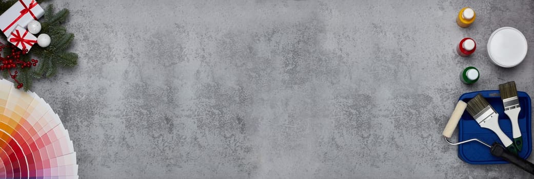 Color palette guide, paint brushes, roller, cans of paint, fir tree branch and gift boxes on grey background. Top view, copy space, New Year and Christmas. Banner background