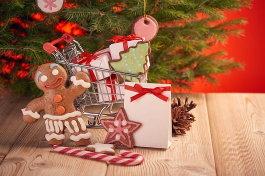 Shopping trolley with gift boxes and gingerbread cookies standing on a wooden table on a red background with Christmas fir tree. Christmas and New Year sale.