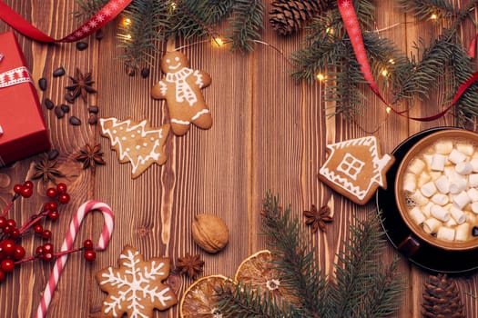 Christmas decor with fir tree branches, glass mug with hot cocoa and marshmallow, gingerbreads and gift box on a wooden background with copy space for text. Top view. For greeting card.