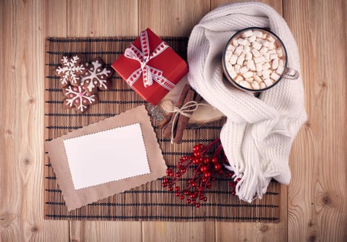 Glass mug with hot cocoa and marshmallow, gingerbread, gift box and blank sheet for text on wooden background. Mockup. Top view. Christmas and New Year greeting card.