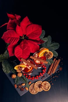 Still life with glasses of mulled wine or fruit tea, Red Poinsettia flowers, fir branch and spices on a dark wooden background. Top view. New year and Christmas table setting.